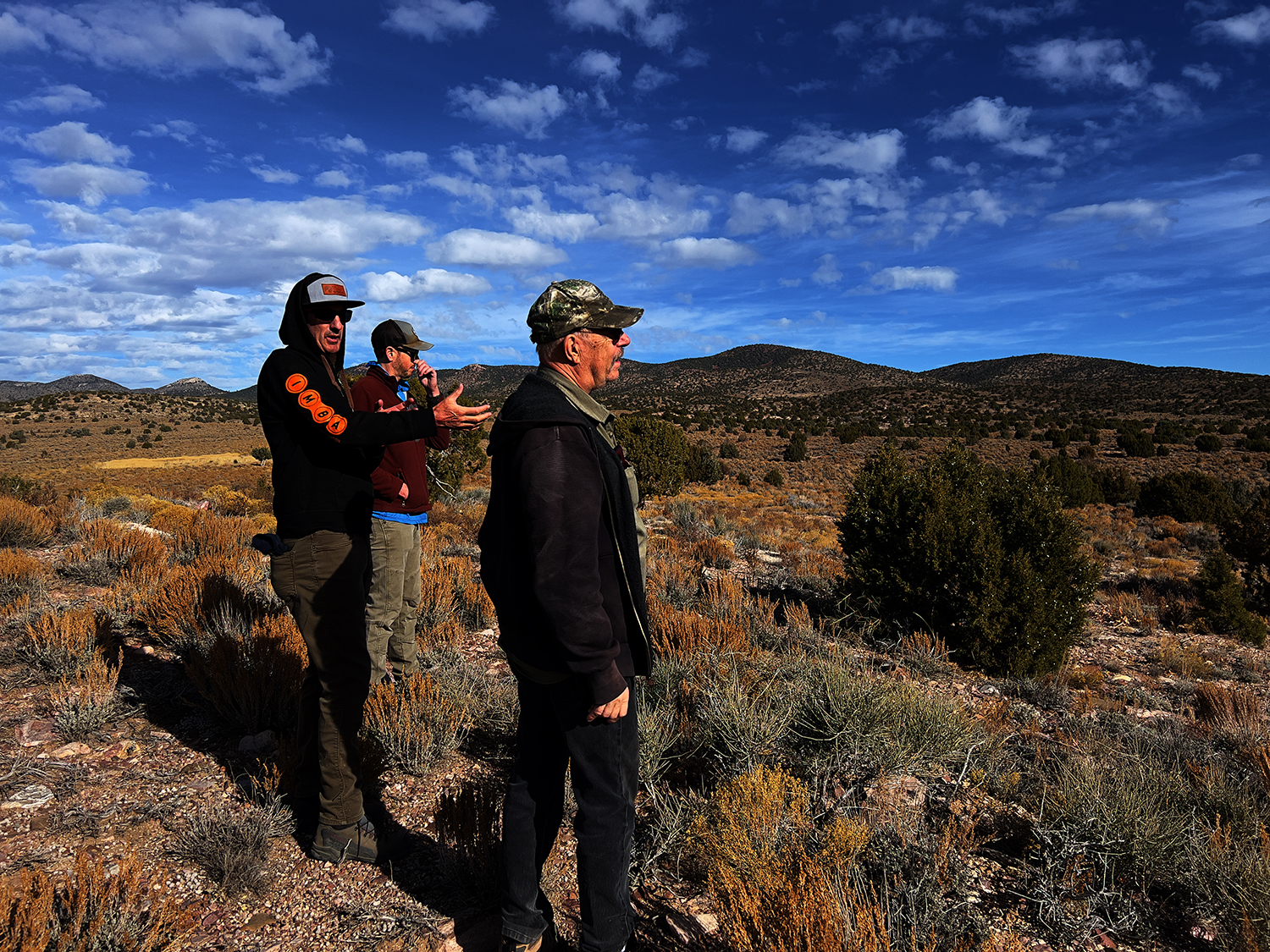 "Three men looking over a desert landscape - man in the middle talking with his hands."