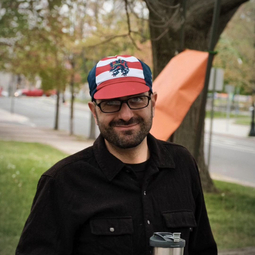 man with black glasses and a short beard wears a cycling cap