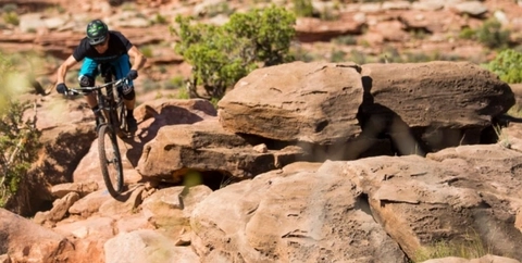 Mountain biking on rugged, rocky BLM Lands in the southwest, a mountain biker rolls down some boulders in blue shorts, knee pads, and a black t-shirt. 