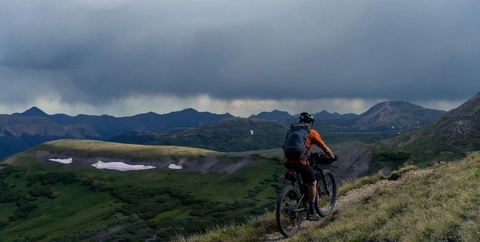 Long-distance bikepacking: a mountain biker rides across dirt singletrack in a green field with a cloudy sky blanketing surrounding mountains.